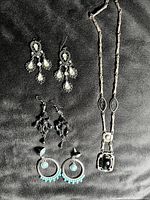 Photo showing three pairs of earrings and one long necklace with black stones on a dark fabric background.