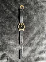 Full view of the watch from the front showing black dial with gold-tone dot and hands, and black leather strap with gold-tone buckle.