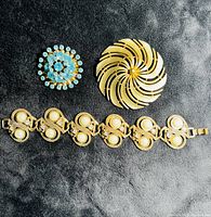 Three vintage jewelry pieces on black fabric background: gold tone swirling Sarah Coventry pin, blue rhinestone brooch, and gold tone bracelet with pearl-like accents.