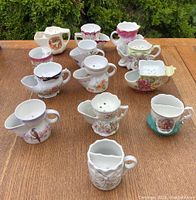 Top down image of 14 assorted porcelain moustache cups on wooden surface showing various floral and decorative patterns, some cups have scalloped edges and guard sides for moustaches.