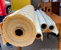 Four rolls of upholstery fabric on a wooden table, showing ends and core of rolls. Yellow and white checkerboard with floral pattern visible on largest roll.