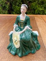 Front view of Royal Doulton porcelain figurine seating a lady in a green historical dress holding a yellow hat.