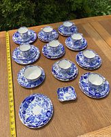 Overall set of nine teacups with matching saucers and various side plates arranged on wooden table with measuring tape to show size.