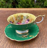Teacup and saucer on a wooden table, showing vibrant green exterior and painted fruit interior of the teacup with gold trim.