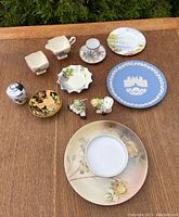 Photo shows 11 assorted china items on a wooden surface including creamers, a sugar shaker, decorative plates, trinket dishes, and clown figurines.