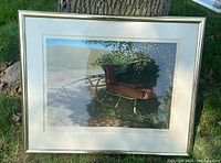 Framed serigraph depicting a wooden cart and greenery with a clear sky background, sitting outdoors against a tree.