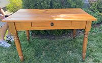 Top and front view of pine table/desk showing rectangular top surface, original drawer with black round knob, and turned legs on grass background.