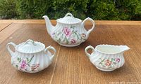 Three-piece Royal Doulton Clovelly pattern set on wooden table showing teapot, creamer, and sugar bowl with floral decoration and scalloped edges.