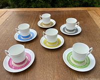 Six coffee cups with matching saucers in different colors arranged on a wooden table.