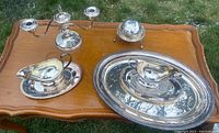 Photo showing all silver-plated items arranged on wooden table outdoors including two candelabras, serving tray, gravy servers, butter dish, small bowl, and ornate plate.