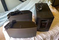 Two black Sonos speakers and Bose subwoofer on a bed, showing the front and side profiles with cords attached