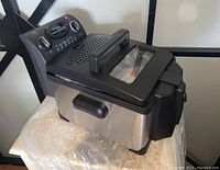 Side view of the T-Fal deep fryer showing stainless steel body, the black handle on lid with window, and control panel with dials and indicator lights.