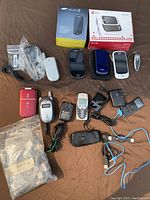 Photo shows a collection of vintage and older model cell phones of different styles and colors, some in original retail boxes, along with chargers, cables, and accessories.