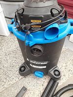 Front view of the MasterVac wet/dry vacuum showing blue top, black body and four caster wheels with hose connectors on the front.
