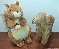Photo shows both plush toys side by side on a wooden surface; left is the brown squirrel holding a small nut, right is a plush camel.