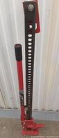 Front view of the red and black ratchet bar jack against a white corrugated wall showing the perforated lifting arm and handle grip.