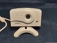 Front view of the off-white COMPRO D-CAM digital camera showing fixed lens, LED indicator, and brand name.