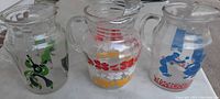 Three clear glass beverage pitchers with various mid-century modern applied color designs, shown from the front angle.