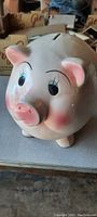 Front view of the vintage ceramic piggy bank showing detailed pig face with blue eyes, pink snout, and pink ears. Visible glazed finish and slight wear marks.