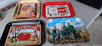 Grouping of four tin serving trays arranged on a surface: Budweiser, Coca-Cola, Beefsteak Tomato, and John Deere designs visible.