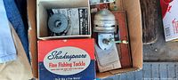 Top-down photo of the lot showing the Shakespeare branded box, original fishing reel, instruction booklet, and paperwork packed inside a cardboard box.