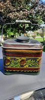 Front view of the miners lunchbox showing floral tole painted design with tin handle and wooden grip