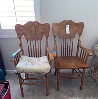 Two wooden dining captain chairs; one has a beige cushion tied to the seat. Both chairs have carved backs with spindle supports and armrests.
