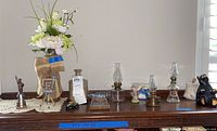 Wide shot showing all collectibles including mini oil lamps, miniature typewriter, car, camera, Statue of Liberty figurine, faux flowers arrangement on burlap, and black bear figurine on a wooden surface.