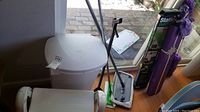 White plastic garbage can with lid alongside mop and dusters in corner of room