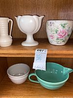 Five pottery items displayed on a wooden shelf including two white vases, a green bowl with pitcher shape, a small grey bowl, and a floral patterned pot.
