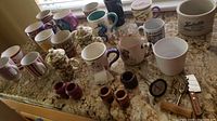 Collection of coffee mugs, ceramic napkin rings, butter crock, and meat thermometer displayed on a granite countertop under a window.