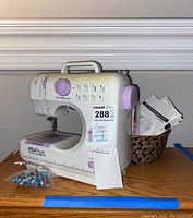Front and side views of Pixie Plus sewing machine with stitch patterns and selector dial visible, instruction manuals in a woven basket behind, and plastic bag of sewing notions in front.