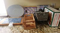 Overview photo showing the group of 8 items including the oval granite trivet, wooden cutting board, black basket, boxed clay baker, boxed kitchen accessories, and wooden kitchen storage box on countertop.