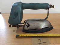 Side profile of the gasoline iron showing metallic base, wooden handle, and fuel tank with temperature adjustment knobs and instructions.