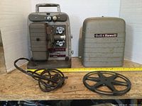 Front and side view of the Bell & Howell 8mm film projector with attached power cord, film reel, and carrying case on wooden surface.