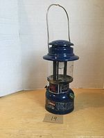 Full view of blue Coleman Deluxe Easi-Lite lantern with metal handle upright, clear glass globe, and metal base with labels.