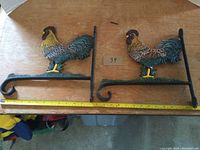 Two painted cast iron rooster mounts shown lying flat with a measuring tape indicating length of about 12.5 inches.