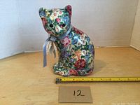 Front view of the porcelain cat figurine with floral chintz pattern, showing the detailed pattern and ribbon bow. A measuring tape indicates the height is about 7.5 inches.