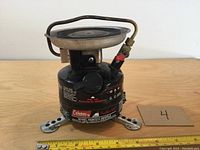 Front view of Coleman mini stove showing brand, model, fuel cap, and legs with measuring tape for scale.