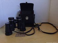 Front view of Taylor binoculars with attached neck strap and black leather case in background on wooden surface.