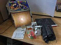 View of the Lite-Brite peg board lit up inside its wood frame showing the peg holes, accompanied by the original box, instruction sheet, plastic bags with colorful pegs, and multiple punched design black cardboard screens stacked on the right.