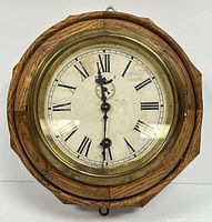 Front view of antique wall clock with octagonal oak frame and brass bezel covering 6-inch dial with Roman numerals.