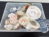 Overhead view of bin containing various cups, saucers, plates, and a pink figurine with assorted floral and scenic patterns.