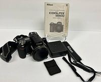 Photo showing Nikon Coolpix 8800 camera, manual, battery charger, and carrying pouch all displayed together on a white surface.