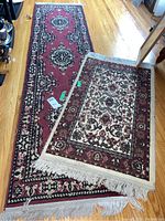 View showing two rugs on hardwood floor: a long 7.5 ft runner with red and patterned design, and a smaller 3x2 cream and red bordered rug with fringes.