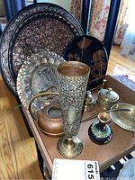 Photo showing three decorative brass plates including one with an Egyptian scene, a tall etched brass vase, small bell, copper watering can, candle snuffer and painted small vase.