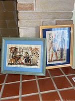 Both framed Egyptian papyrus prints placed against a stone wall and tile floor, showing overall size and framing.