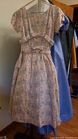 Photo of a peach and blue floral embroidered dress with short puff sleeves, fitted waist with two bow accents, pleated knee-length skirt, hanging on a hanger. Purple satin dress is visible behind.