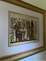 Framed Egyptian papyrus artwork with gold decorative frame and glass front, angled view showing artwork details and hieroglyphics.