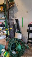 Wide shot showing rolled wire fencing, metal tomato cages, green metal stakes, garden hose, and watering container